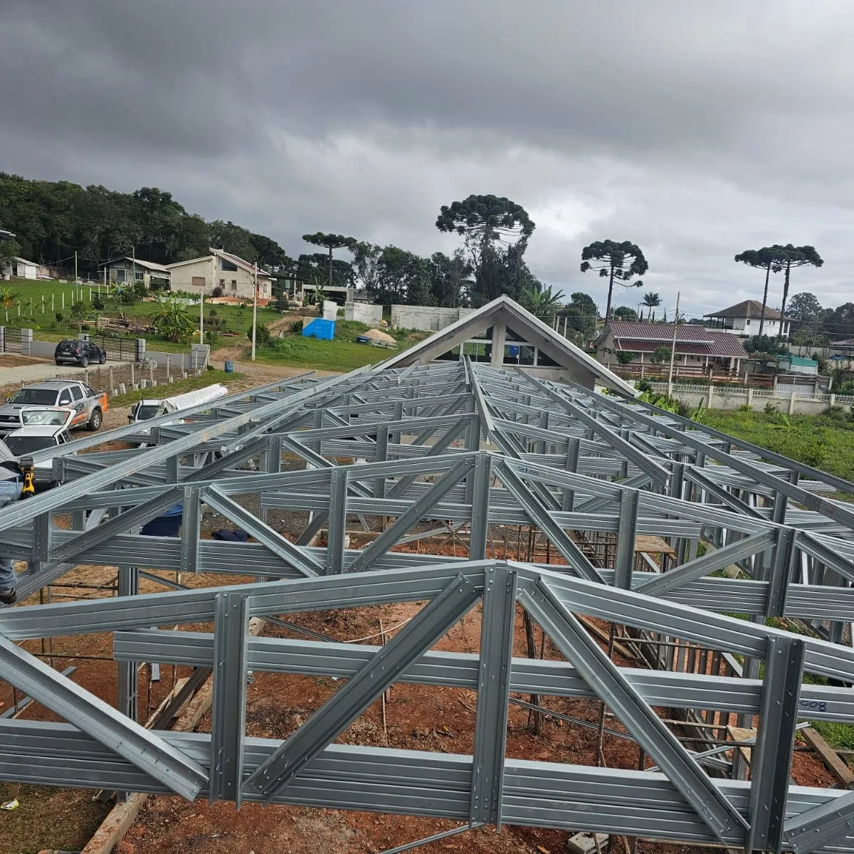 Treliças de aço galvanizado da cobertura montadas sobre a obra, com araucárias e casas ao fundo.