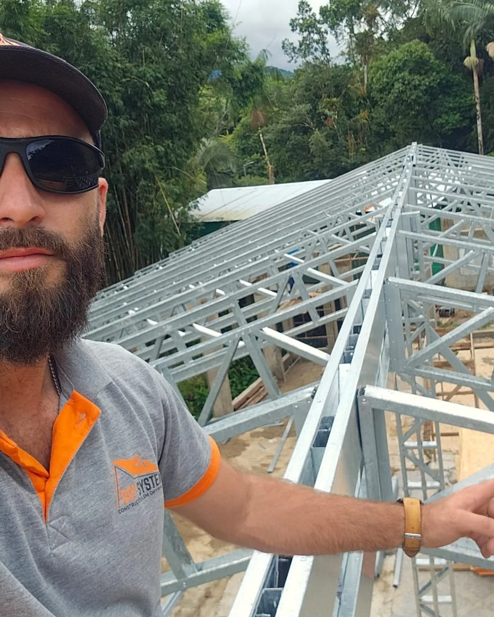 Integrante da equipe, de boné e camisa da empresa, apontando para as treliças da cobertura.