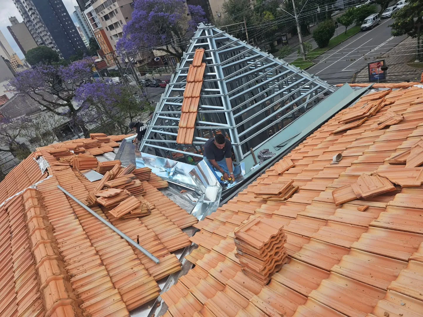Estrutura metálica em pirâmide sobre um telhado, com telhas cerâmicas sendo assentadas.